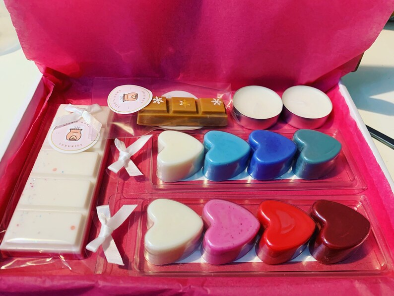 May include: A pink gift box filled with scented wax melts. The box contains a variety of heart-shaped wax melts in blue, red, pink, and white. There are also two small white tea lights and a bar of wax melt in a brown wrapper.