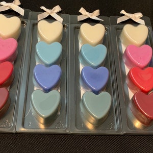May include: Four sets of heart-shaped wax melts in various colors, including white, pink, red, blue, and purple. Each set is packaged in a clear plastic container with a white ribbon.