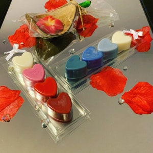 May include: A set of six heart-shaped wax melts in a clear plastic packaging. The wax melts are in shades of red, pink, blue, and white. The packaging is on a reflective surface with red rose petals.