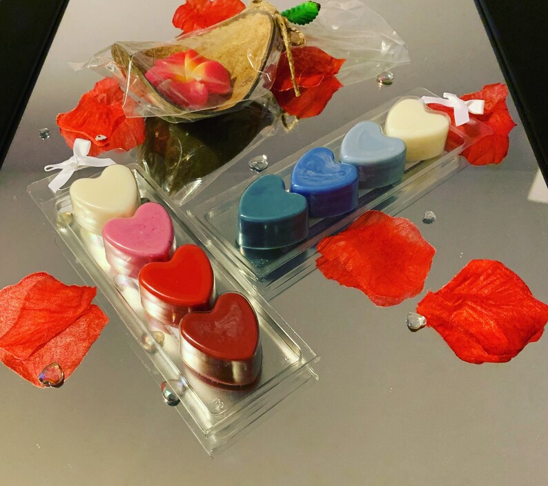 May include: A set of six heart-shaped wax melts in a clear plastic packaging. The wax melts are in shades of red, pink, blue, and white. The packaging is on a reflective surface with red rose petals.