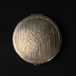 May include: A silver compact with an engraved design of two figures in a garden setting. The compact has a raised border and a blank rectangular space for engraving.