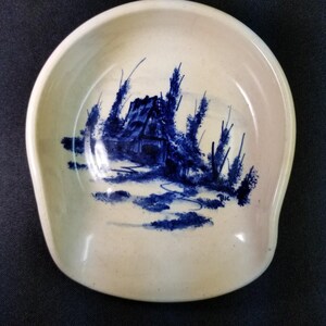 May include: Off-white ceramic spoon rest with a blue landscape design. The design features a house and trees. The spoon rest has a curved shape with a raised edge. The design is hand-painted.