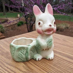May include: White ceramic bunny planter with green leaves and a pink nose. The planter has a small pot for planting a small plant.