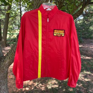 May include: Red vintage jacket with a yellow vertical stripe and a zip-up front. The jacket features a Cooper patch on the chest. The jacket has a classic, retro style.