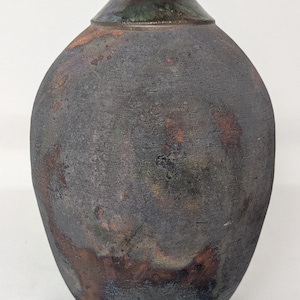 May include: A handcrafted ceramic vase with a dark, textured exterior. The vase has a rounded body and a narrow neck with a dark green and maroon glaze. The overall aesthetic is rustic and earthy, suitable for home decor.