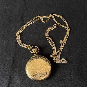 May include: A gold-colored pocket watch with an ornate floral design on the cover, suspended from a matching gold chain. The watch and chain are set against a black background, creating a striking contrast.