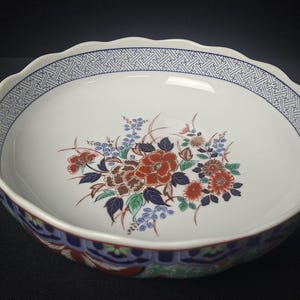 May include: A white ceramic serving bowl with a scalloped edge and floral design. The bowl features a blue geometric border and a central arrangement of red, brown, and blue flowers and leaves.