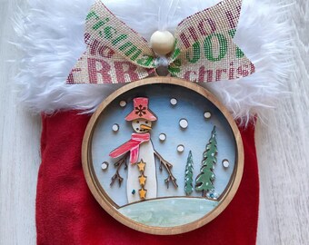 May include: A wooden snowman ornament with a red hat, scarf, and buttons. The ornament is surrounded by a blue background with white snowflakes and two green pine trees. The ornament is hanging from a red stocking with a white fur cuff and a burlap bow with the text "Merry Christmas".