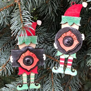 以下が含まれることがあります： 赤い帽子と緑の帽子をかぶった木製のエルフのオーナメント2つが、「Elf Cam」と書かれたカメラを持っています。オーナメントは緑色の松の木にぶら下がっています。