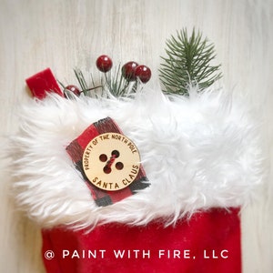 Puede incluir: Una media roja y blanca con un puño de piel blanca. Un botón de madera con el texto "Property of the North Pole Santa Claus" está unido al puño.