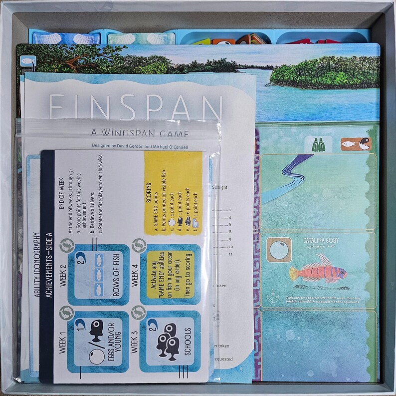 May include: A Finspan board game with a blue and green color scheme. The game board features a variety of fish illustrations and the text "Finspan: A Wingspan Game". The game board is divided into four sections, each with a different week of the game. The game board also includes a scoring section with a list of scoring criteria.