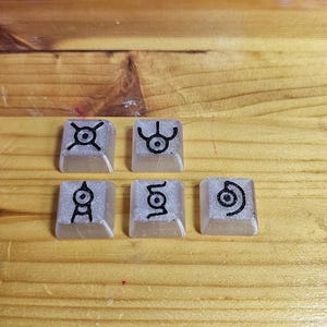 以下が含まれることがあります： 半透明の銀色のグリッターの背景に、ユニークな黒いシンボルが付いた5つのカスタムキーキャップ。キーキャップは正方形で、木製の表面に置かれており、背景にはキーボードが見えます。