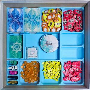 May include: A blue and white game board with various compartments for storing game pieces. The compartments contain small, colorful plastic pieces, including fish, gears, and other shapes. The board also includes a card that says "Consumed Fish" with a number 2 on it.