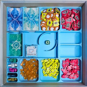 May include: A blue organizer with compartments for storing game pieces. The compartments contain various colored plastic pieces, including yellow fish, red fish, and orange spheres. The organizer also has a compartment labeled "Consumed Fish".