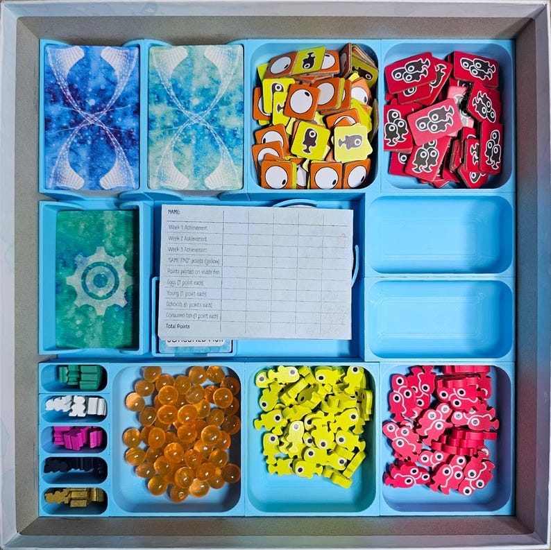 May include: A blue game board with compartments for storing game pieces. The compartments contain various colored plastic pieces, including red, yellow, orange, and blue. The board also has a score sheet with the title "NAME:  Week 1 Achievement:  Week 2 Achievement:  SAVE 100 points (glow) Points earned on each roll:  Eggs (2 point each)  Fish (3 point each)  Octopus (6 point each)  Bonus (if earn 6 point each)  Total Points"