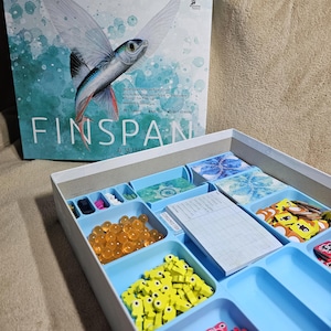 May include: The game box for Finspan: A Winged Game, featuring a blue and green illustration of a flying fish. The box is open, revealing a blue tray with compartments for game pieces, including orange beads, yellow fish, red fish, and brown fish.