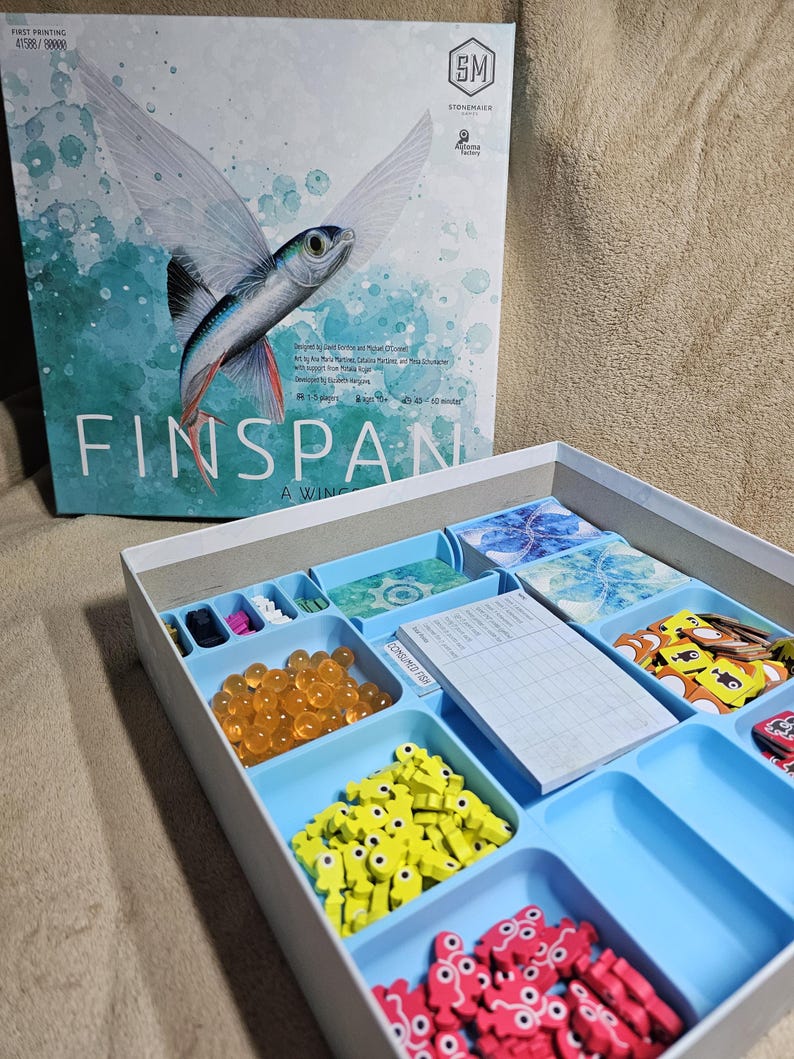 May include: The game box for Finspan: A Winged Game, featuring a blue and green illustration of a flying fish. The box is open, revealing a blue tray with compartments for game pieces, including orange beads, yellow fish, red fish, and brown fish.