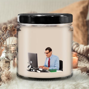 May include: A white candle jar with a black lid. The jar has a picture of a person sitting at a desk and using a computer. The person is wearing glasses and a blue shirt with a red tie.