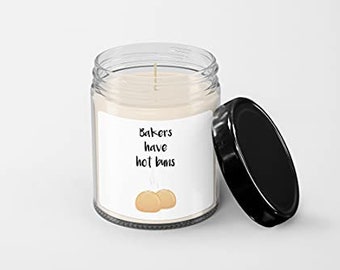 May include: A clear glass candle jar with a black lid. The candle is white and has a label that reads "Bakers have hot buns" with a cartoon image of two buns.