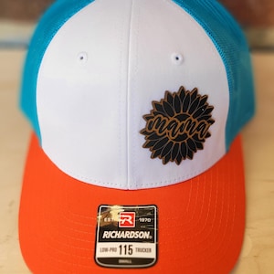 May include: A trucker hat with a white front panel, a blue mesh back, and an orange brim. The hat features a sunflower design with the word "mama" and a small orange button on top.