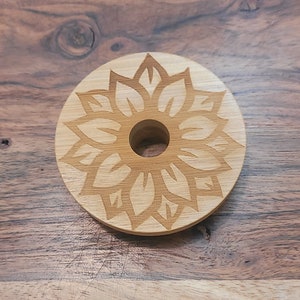 May include: A round wooden coaster with a laser-etched mandala design. The coaster is light brown and has a hole in the center.