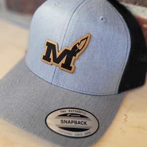 May include: A light blue and black baseball cap with a brown wooden patch featuring a black letter "M" and a feather design. The cap has a black mesh back and a gray front panel. The cap has a silver label that reads "The Authentic Yupoong Snapback Yupoong Since 1974."