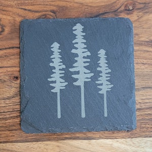 May include: A square, dark gray slate coaster with a light gray etched design of three stylized pine trees. The coaster has a natural, rough edge and sits on a wooden surface.
