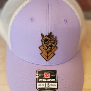 May include: A lavender trucker hat with a white mesh back. The hat has a wooden patch with a floral design and the text "Richardson" and "Low-Pro 115 Trucker" on the front.