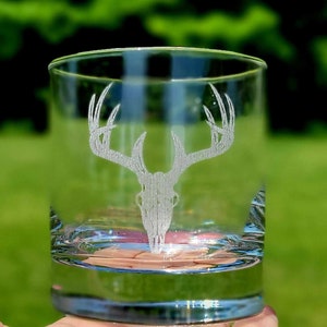 May include: A clear glass tumbler with an etched design of a deer skull with antlers. The glass is held in a hand.