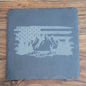 May include: A gray slate coaster with a white design of a mountain range and an American flag.