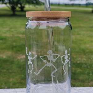 May include: A clear glass drinking tumbler with a bamboo lid and a glass straw. The tumbler has an etched design of three skeletons dancing.