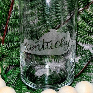 May include: A clear glass can with an etched design of the state of Kentucky and the word "Kentucky" on it. The can is sitting on a gray surface with green foliage around it.
