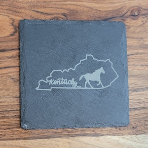 May include: A black slate coaster with an engraved outline of the state of Kentucky and a running horse. The word "Kentucky" is engraved below the horse.
