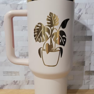 May include: A pink stainless steel travel mug with a gold lid and a gold graphic of a potted plant with leaves.