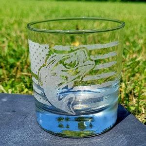 May include: A clear glass tumbler with an etched design of a bass fish and an American flag. The glass is sitting on a dark gray surface.