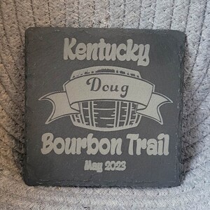 May include: Black slate coaster with a white design of a barrel with a banner that says "Doug" and the text "Kentucky Bourbon Trail May 2023".