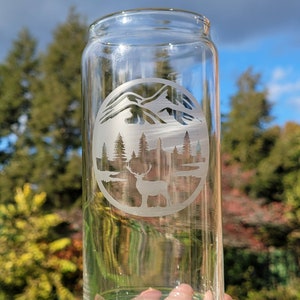 May include: A clear glass drinking glass with an etched design of a mountain range, trees, and a deer in a circle.