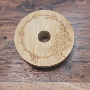 May include: A round, light brown bamboo coaster with a floral design engraved around the edge. The coaster has a hole in the center.