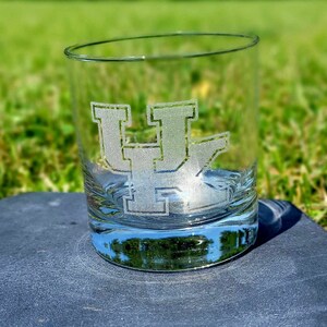 May include: A clear glass tumbler with a frosted etching of the letter "UK" on the front. The tumbler is sitting on a gray surface.