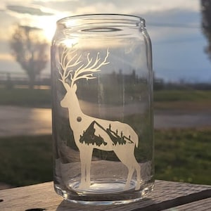 May include: A clear glass jar with a white silhouette of a deer with antlers and a mountain range inside. The jar is sitting on a wooden surface.