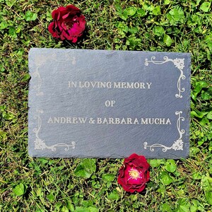 May include: A black slate plaque with a white inscription that reads "In loving memory of Andrew & Barbara Mucha". The plaque is surrounded by green grass and two red roses.