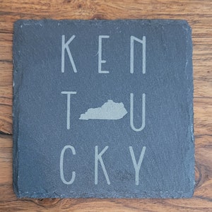 May include: A black slate coaster with the word "KENTUCKY" spelled out in white letters. The state of Kentucky is also depicted in white in the center of the coaster.