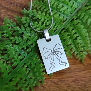 May include: A silver necklace with a rectangular pendant featuring a simple line drawing of a bow. The pendant is hanging on a silver chain.