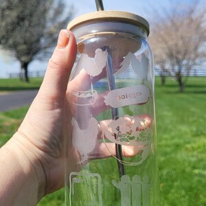 May include: A clear glass tumbler with etched food and icon designs, including a chicken, hot dog, and computer. It has a bamboo lid and a metal straw. The tumbler is held in front of a blurred outdoor background.