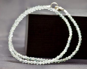 May include: A delicate necklace composed of small, pale green beads. The necklace is strung together with a silver clasp. The beads are evenly spaced, creating a subtle, elegant design. The necklace is displayed against a dark background.