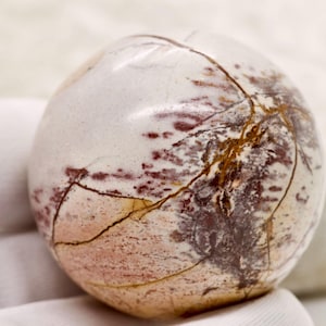 Pode incluir: Uma pedra esférica polida, com uma base creme, acentuada por veios castanho-avermelhados e dourados. A superfície da pedra tem um acabamento liso e brilhante. É segurada por uma luva branca. Esta pedra decorativa é um item de decoração único.