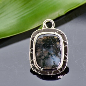 Puede incluir: Un colgante de plata con una piedra de ágata musgosa de forma cuadrada. La piedra tiene un patrón verde y gris.