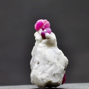 Könnte beinhalten: Eine Gruppe von rosafarbenen Rubinkristallen auf einem weißen Stein. Die Rubinkristalle sind zusammen gruppiert und haben ein glänzendes, durchscheinendes Aussehen.