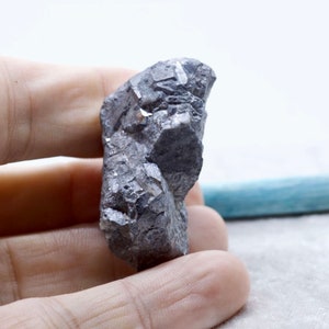 May include: A rough, gray, metallic rock with a crystalline structure. The rock is about 2 inches tall and is held in a hand.