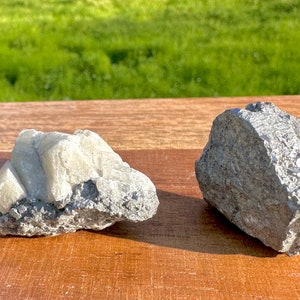 Puede incluir: Dos rocas grises ásperas con cristales blancos. La roca de la izquierda tiene un grupo de cristales blancos en la parte superior. La roca de la derecha tiene algunos cristales blancos más pequeños dispersos.